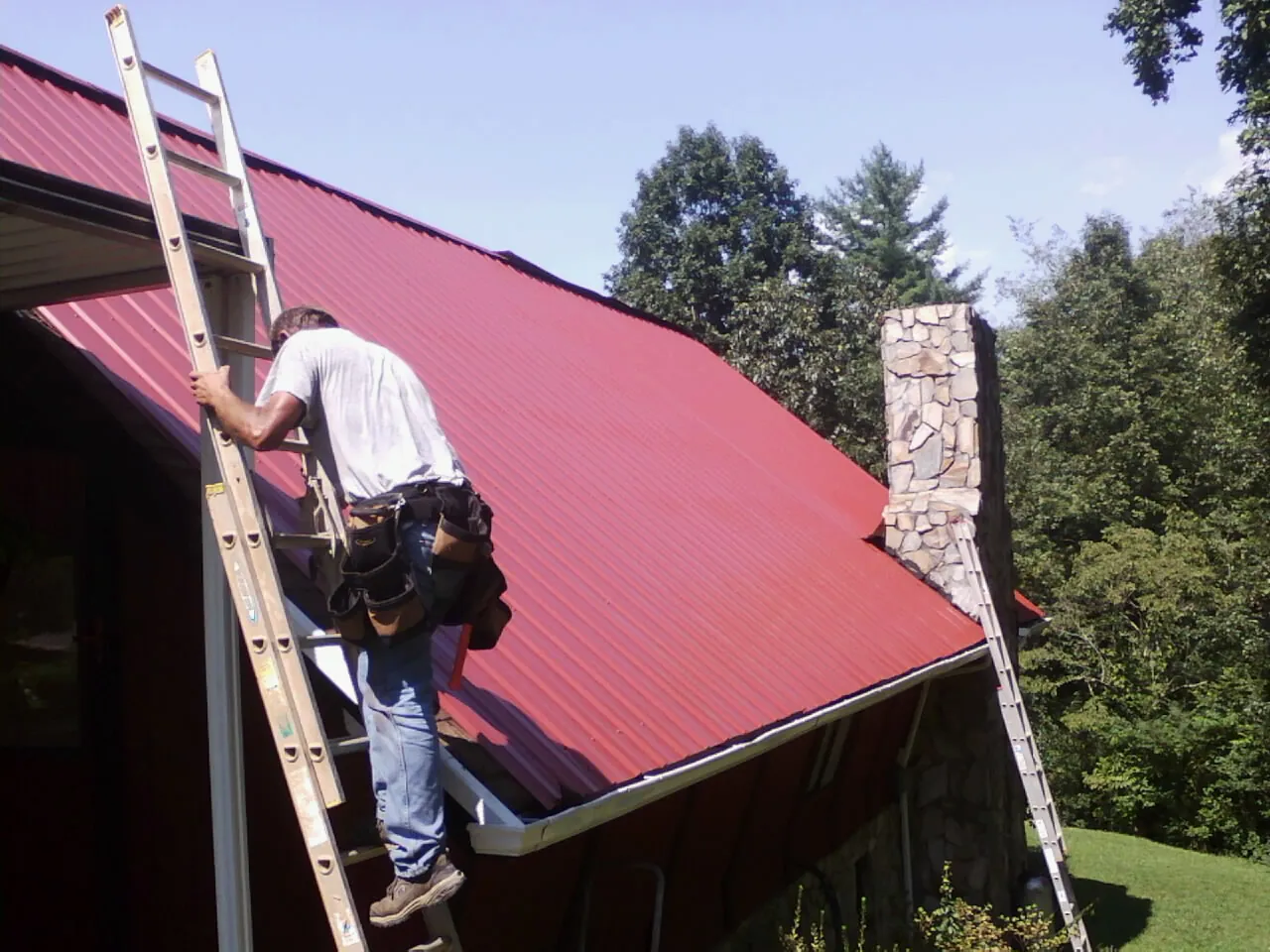 Roofer on a ladder beside a red metal roof and stone chimney — Soffit & Fascia Repair by Roofing Brothers in Waterboro