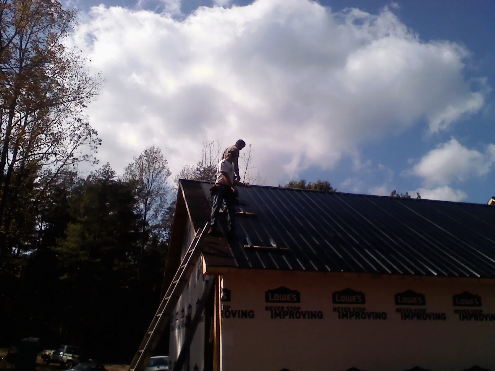 Roofer fastening dark metal panels high against a cloudy sky, from a Roof Inspection job in Waterboro